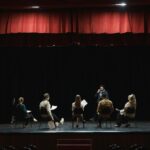 A group of actors rehearsing on a dimly lit stage in an auditorium, preparing for a theater performance.